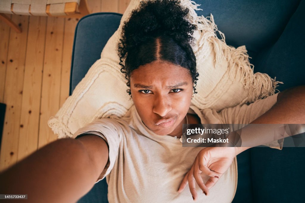Portrait of girl puckering lips while taking selfie lying on sofa at home