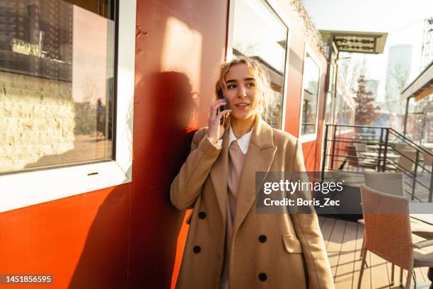 schönes blondes mädchen telefoniert an einem sonnigen tag mit einem mobiltelefon, während es sich an eine rote wand lehnt - portraits fun mobile wall stock-fotos und bilder