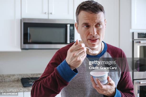 man eats a cup of yogurt while standing in kitchen - yogurt stock pictures, royalty-free photos & images