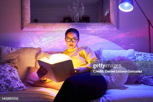 woman in glasses reading an illuminated book while relaxing on a sofa at home. - fantasy stock-fotos und bilder