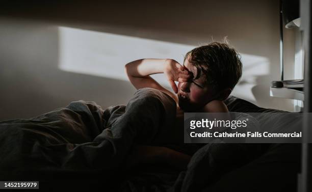 a little boys grimaces as the daylight disturbs his sleep, he covers his eyes - boy asleep in bed stock-fotos und bilder