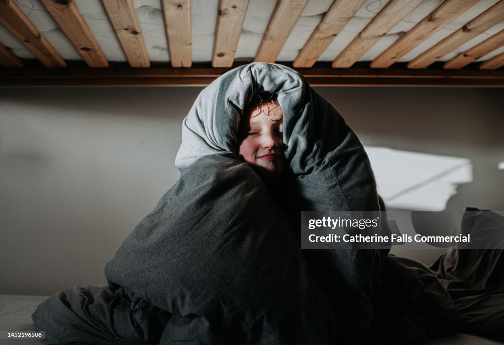 A child, wrapped in a grey duvet, peers out and smiles at the camera