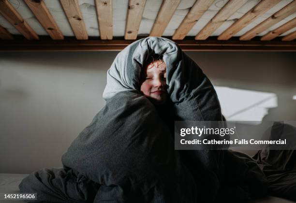 a child, wrapped in a grey duvet, peers out and smiles at the camera - linge de lit photos et images de collection