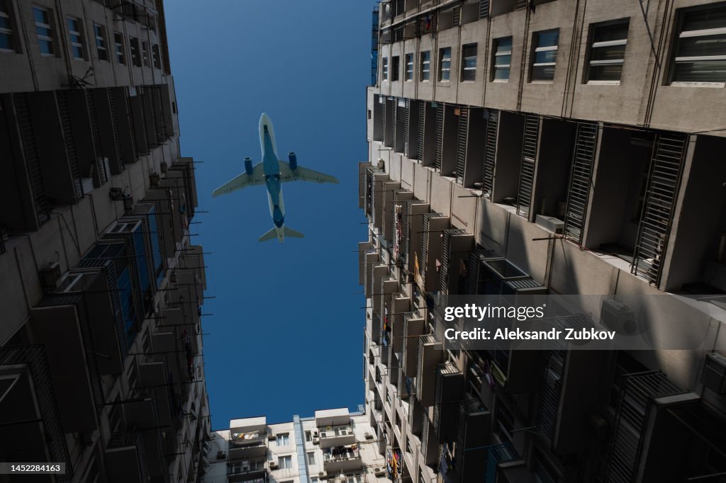 A passenger plane flies low over a house or building in a residential area. Bottom view. The plane is on the background of a blue cloudless sky, landing or taking off near the city. Tourist destinations.