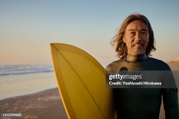 smiling senior holds surfboard and stands next to rising sun - ミッドアダルト ストックフォトと画像