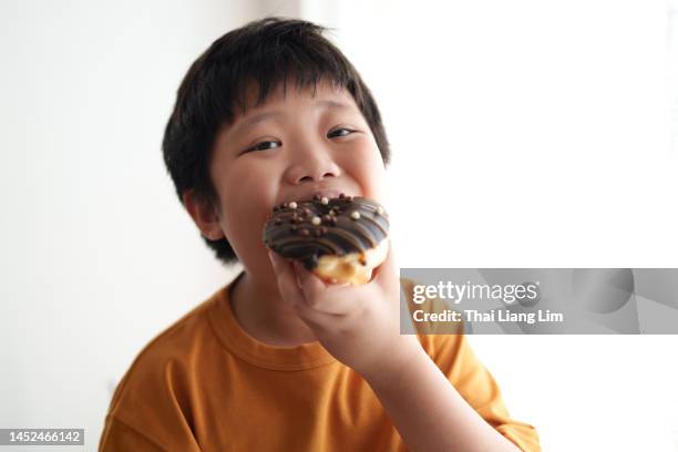 asian boy biting into a chocolate doughnut - eating donuts stockfoto's en -beelden