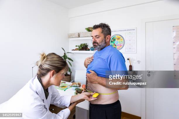 female nutricionist doctor in her office in a medical consultation mesuring corporal body mass index and waist contour with a mesuring tape off an overweight  male patient - body mass index chart stock pictures, royalty-free photos & images