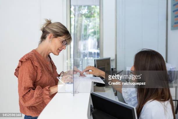 mujer joven está llenando documentos en la recepción de una clínica médica y tomando con recepcionista - receptionist fotografías e imágenes de stock