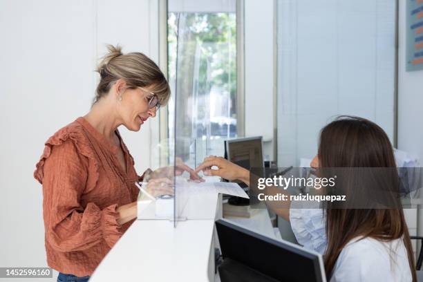 young woman is filling documents at a medical clinic reception and taking with female receptionist - reception desk stock pictures, royalty-free photos & images