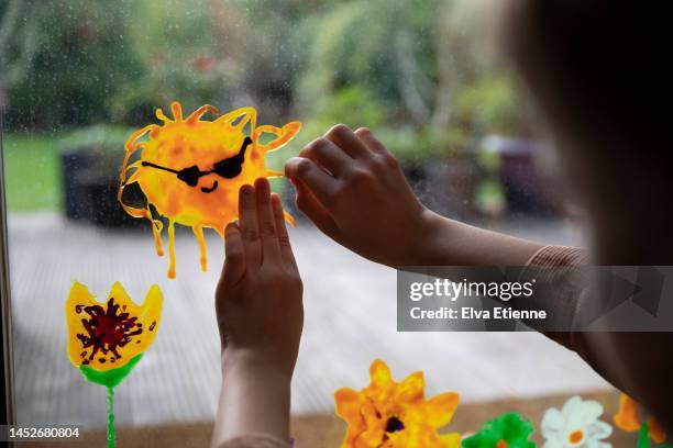 child arranging a smiling sunshine sticker created from window paint, onto a large glass window looking out onto a back yard. - girls hands behind back stock-fotos und bilder