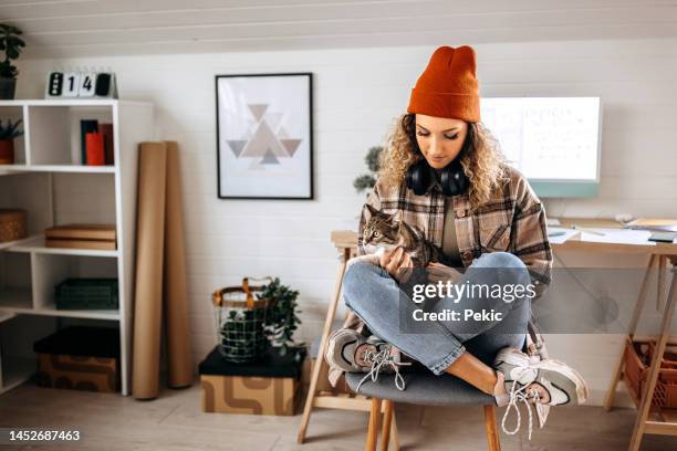 joven y hermosa estudiante que se une a su gato mientras se toma un descanso de estudiar en su acogedor apartamento - cat with red hat fotografías e imágenes de stock