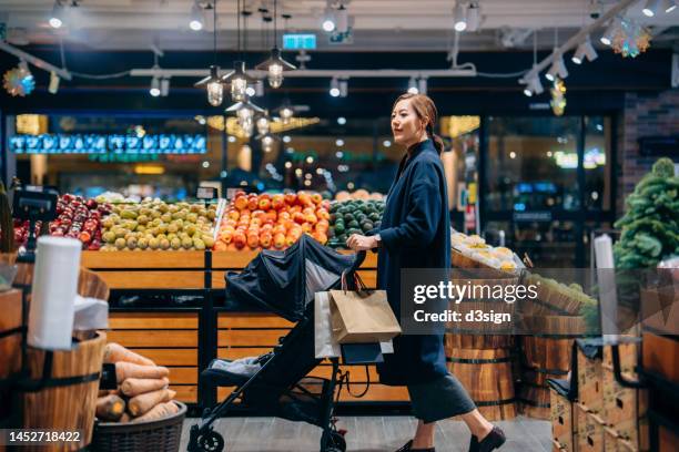 young asian mother pushing a baby stroller, grocery shopping with her baby in supermarket. walking along fruit and vegetables aisle. routine shopping. healthy eating lifestyle - produce aisle photos et images de collection