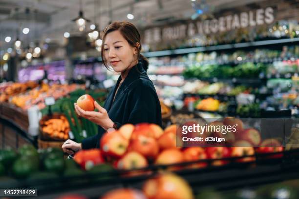 young asian woman grocery shopping for fresh organic fruits in supermarket, choosing red apple along the produce aisle. routine grocery shopping. healthy eating lifestyle - 生鮮食品コーナー ストックフォトと画像