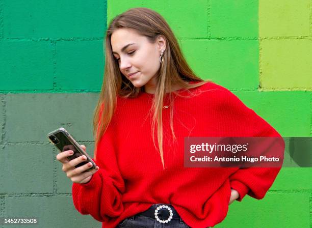 teenage blonde girl with a red sweater surfing with her mobile phone. green wall background - portraits fun mobile wall stock-fotos und bilder