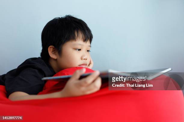 boy lying down on a bean bag and holding a book. children leisure reading concept - boy lying down stock pictures, royalty-free photos & images