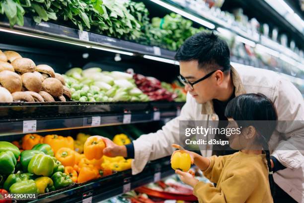 young asian father and her little daughter grocery shopping in supermarket. they are choosing fresh organic bell peppers together along the produce aisle. fruits and vegetables shopping. routine grocery shopping. going green and healthy eating lifestyle - supermarket fruit stockfoto's en -beelden