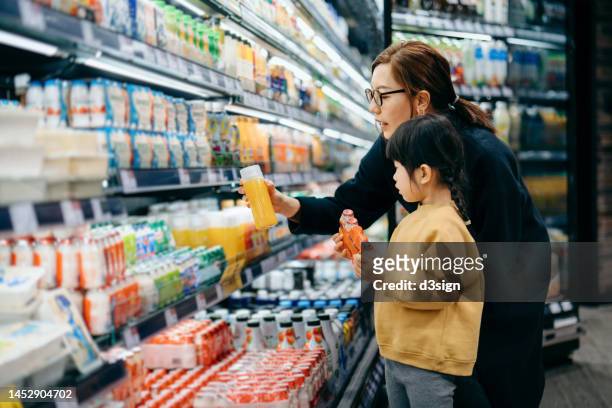 young asian mother and her little daughter grocery shopping in supermarket. they are choosing fresh fruit juice together along the beverage aisle. routine grocery shopping. healthy eating lifestyle - child stock photos et images de collection
