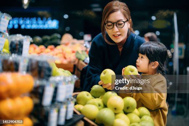 young asian mother and her little daughter grocery shopping in supermarket. they are choosing fresh organic green apples together along the produce aisle. fruits and vegetables shopping. routine grocery shopping. going green and healthy eating lifestyle - family shopping list stock pictures, royalty-free photos & images