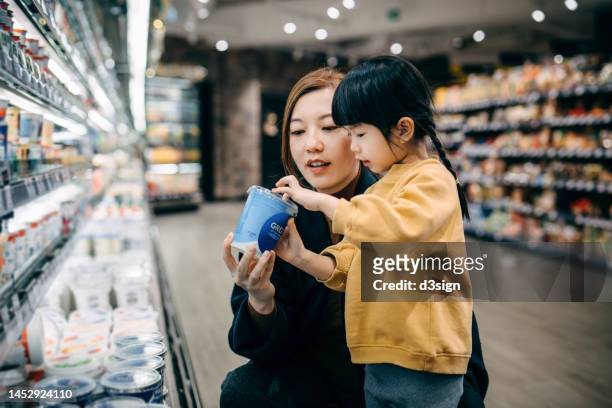 young asian mother and her little daughter grocery shopping in supermarket, choosing dairy product along the dairy aisle. reading the nutrition label on a container of organic healthy yoghurt. healthy eating lifestyle - tag 2 fotografías e imágenes de stock