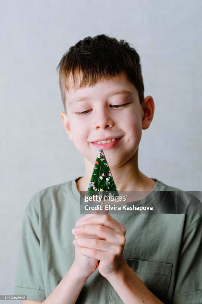 A boy in a green T-shirt sucks a lollipop in the form of a Christmas tree, against a white wall
