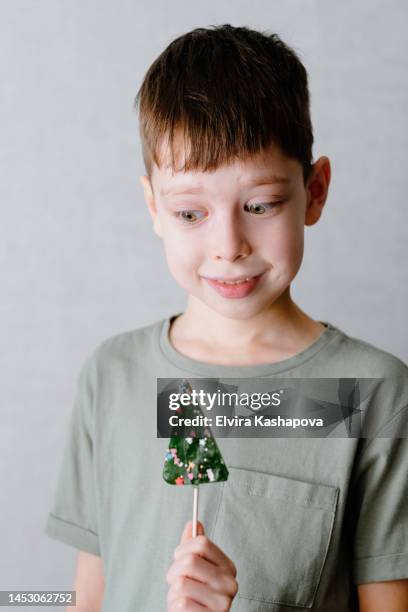 A Boy In A Green Tshirt Sucks A Lollipop In The Form Of A Christmas Tree Against A White Wall High-Res Stock Photo