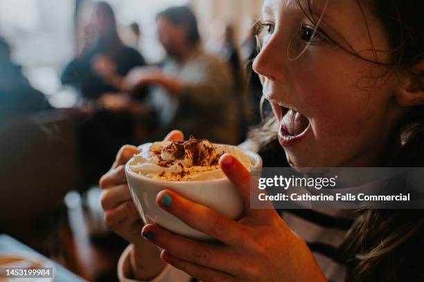 a little girl shelters from the harsh weather, inside a coffee shop. she takes a first sip from her hot cocoa. - saucer stock-fotos und bilder