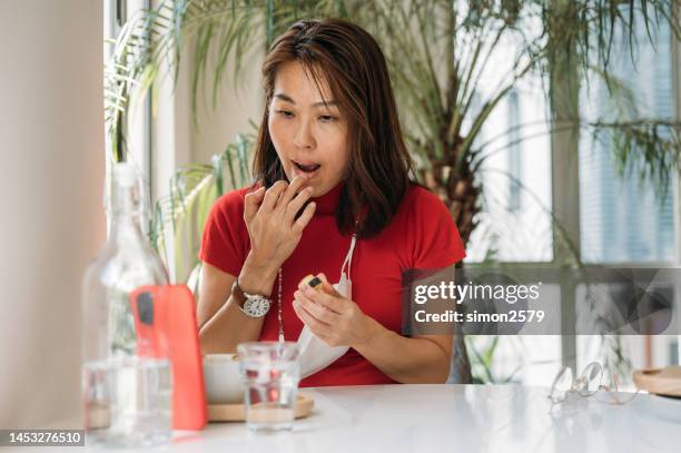 eine asiatische frau, die lippenbalsam aufträgt und das smartphone als spiegel im co-working space benutzt - lip balm stock-fotos und bilder