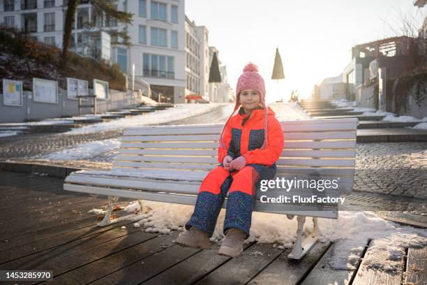 caucasian girl in warm clothing sitting on a bench in the city, during sunny winter day - moon boot stock pictures, royalty-free photos & images