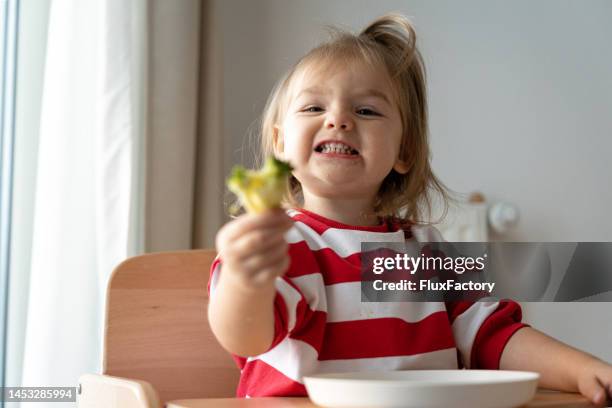 toddler girl holding broccoli, while siting in high chair - steamed vegetables stock pictures, royalty-free photos & images