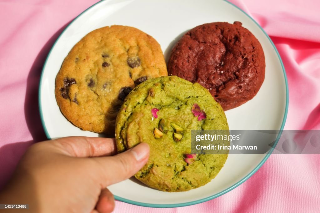 Picking up cookie, a plate filled with pistachio cookie, red velvet cookie, choco chip cookie