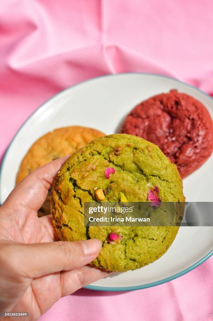 Picking up cookie, a plate filled with pistachio cookie, red velvet cookie, choco chip cookie