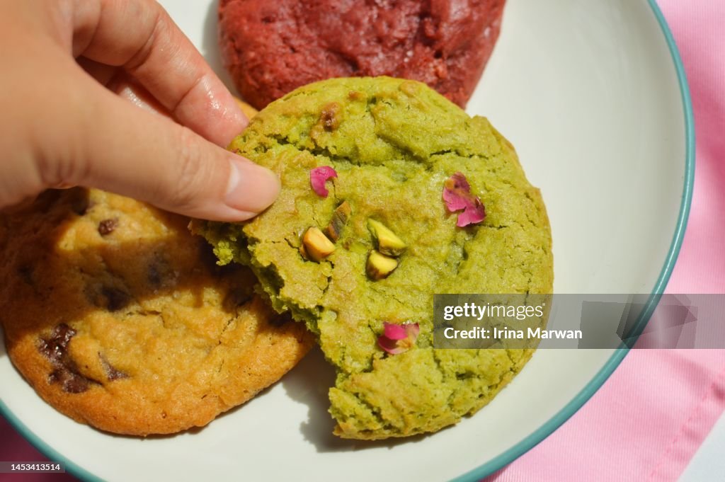 Picking up cookie, a plate filled with pistachio cookie, red velvet cookie, choco chip cookie