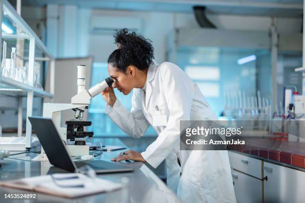 wissenschaftlerin, die unter dem mikroskop schaut und einen laptop in einem labor benutzt - forschung labor stock-fotos und bilder