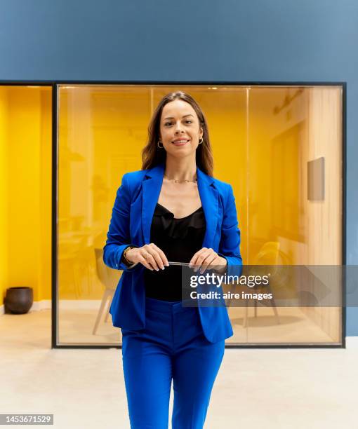 young smiling long-haired woman elegantly dressed holding a pen in her hand standing looking at the camera inside her office - yellow suit stock pictures, royalty-free photos & images