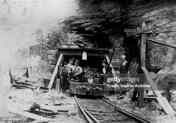 View of coal miners, including a young boy, next to a mine cart at a drift mine entrance in West Virginia, 1908. An exposed live wire, set at...