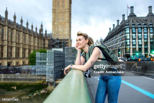 attractive caucasian young adult female tourist leaning on fence of the bridge over thames in london and adoring the view - railing stockfoto's en -beelden