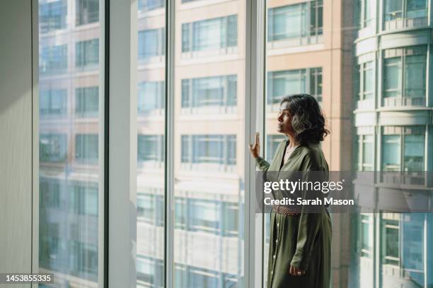 businesswoman standing with hand against office window looking out - sarah green stock pictures, royalty-free photos & images