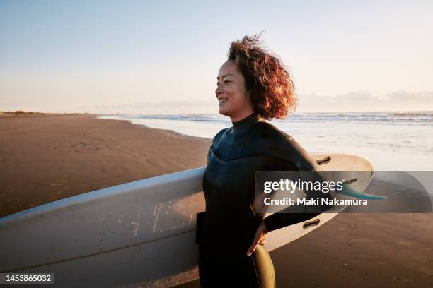 side view portrait of a woman standing in the surf with a surfboard under her arm. - lifestyle imagens e fotografias de stock