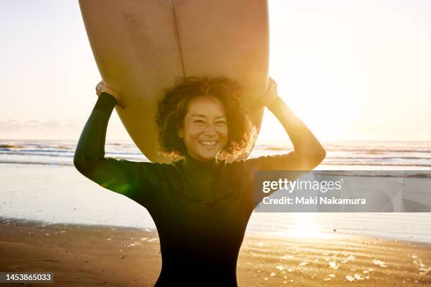 portrait of a curly-haired woman smiling with a surfboard on her head. - arms raised sunrise stock pictures, royalty-free photos & images