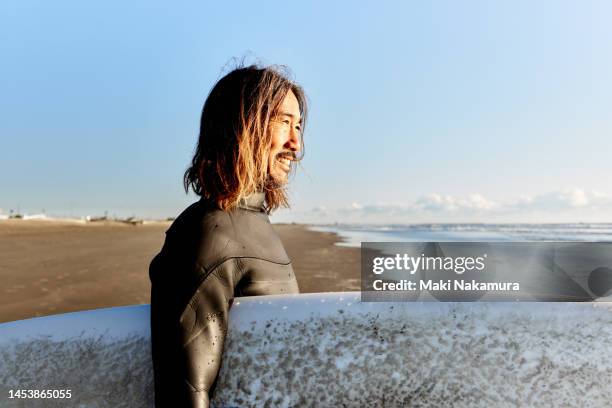 side view portrait of a man standing in the sun with a surfboard under his arm, his face dazzled by the sun's rays. - forte beach photos et images de collection