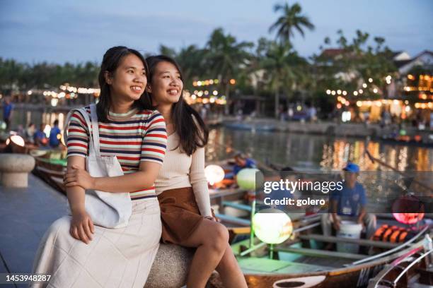 portrait of two young asian woman sitting at riverside in hoi an at sunset - vietnamese culture stock pictures, royalty-free photos & images