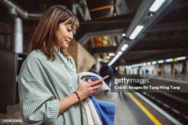 pretty asian woman using smartphone while waiting on railway station platform - day in the life fotografías e imágenes de stock