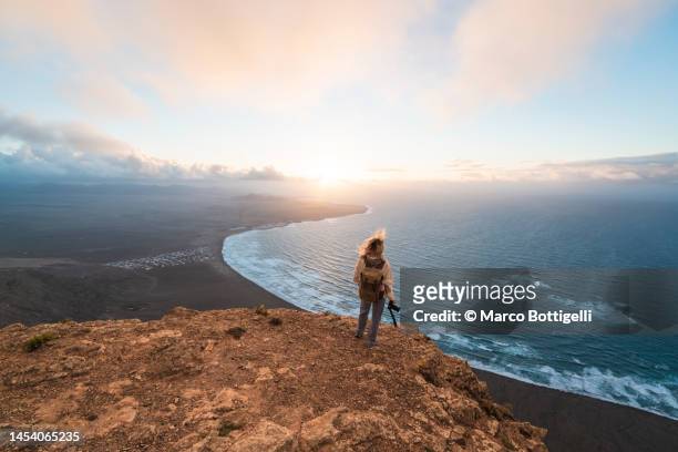 woman with camera looking at view from the top of a cliff at sunset in lanzarote, spain - ocean cliff stock pictures, royalty-free photos & images
