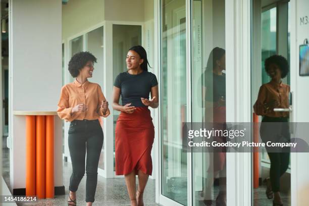 diverse young businesswomen talking while walking along a hallway at work - women side by side stock pictures, royalty-free photos & images