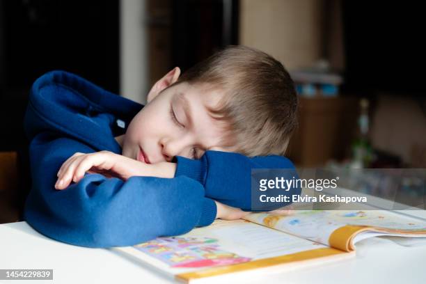 a boy of 8 years old in a hoodie fell asleep reading a book at the table. child doing boring homework at home - 8 9 years stock pictures, royalty-free photos & images