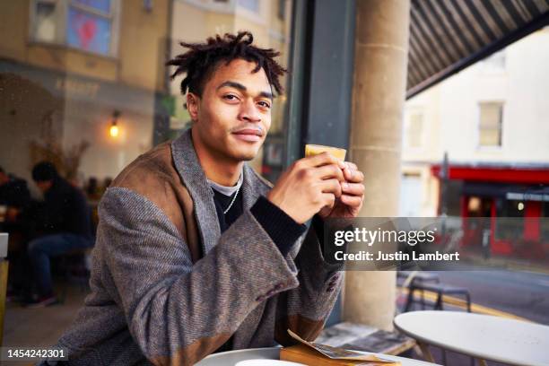 fashionable young man smiling and looking to camera while enjoying a cup of coffee outside a cafe - sidewalk cafe stock pictures, royalty-free photos & images