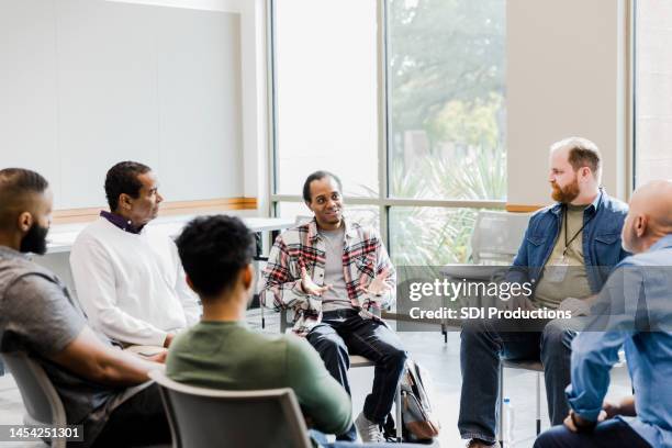 hombres diversos escuchan a joven compartiendo en reunión de terapia - terapia de grupo fotografías e imágenes de stock