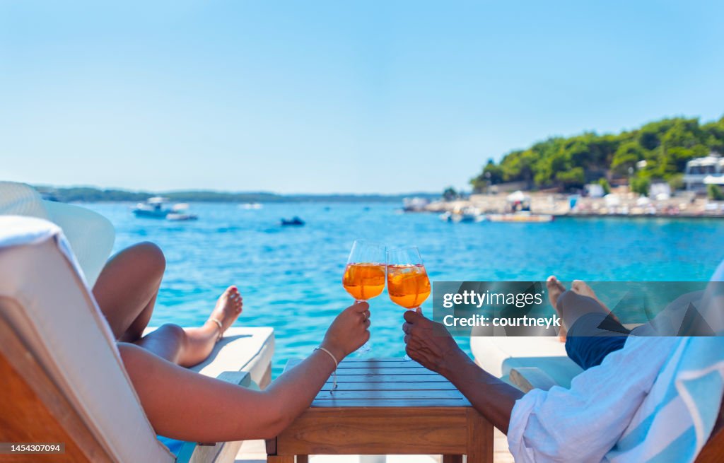 Paar entspannt und toastet mit einem Aperol Spritz Cocktail auf einem Stranddeck über dem Meer.