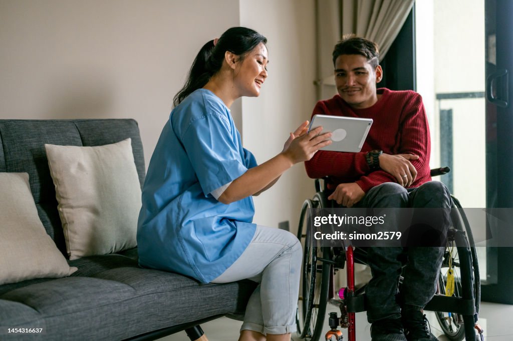 An Asian female healthcare worker teaching Mature Asian disabled Malay man in wheelchair how to online by using digital table at living room.