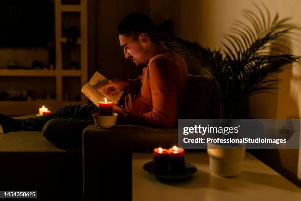 während einer energetischen krise liest ein mann im dunkeln ein buch mit brennenden kerzen. - kerzenschein stock-fotos und bilder